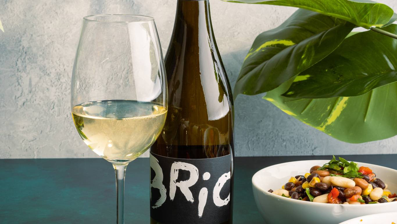 Bottle of Brio Pinot Grigio available at Virgin Wines, which is vegan, stood next to a glass of white wine, a bean salad bowl and a plant