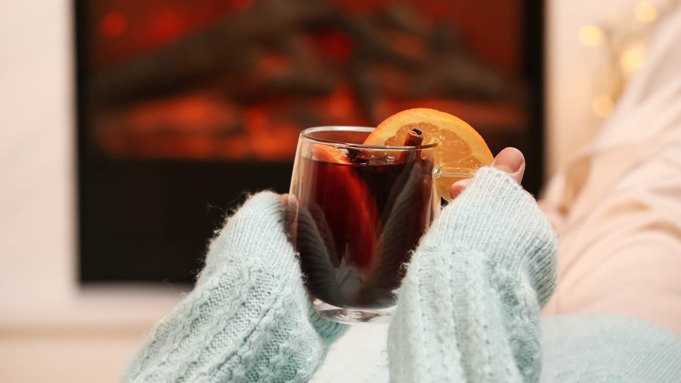 Woman in fluffy blue jumper holding a glass mug of mulled wine on a sofa by the fireplace