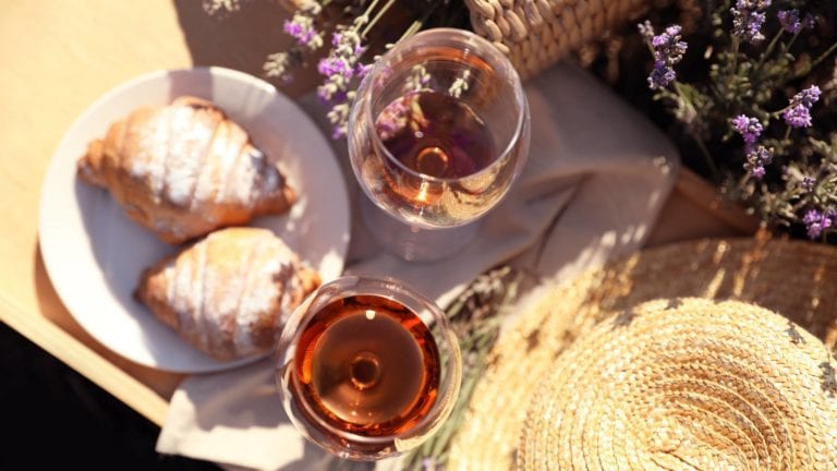 Plate with croissants and glasses of wine on wooden tray in lavender field, top view