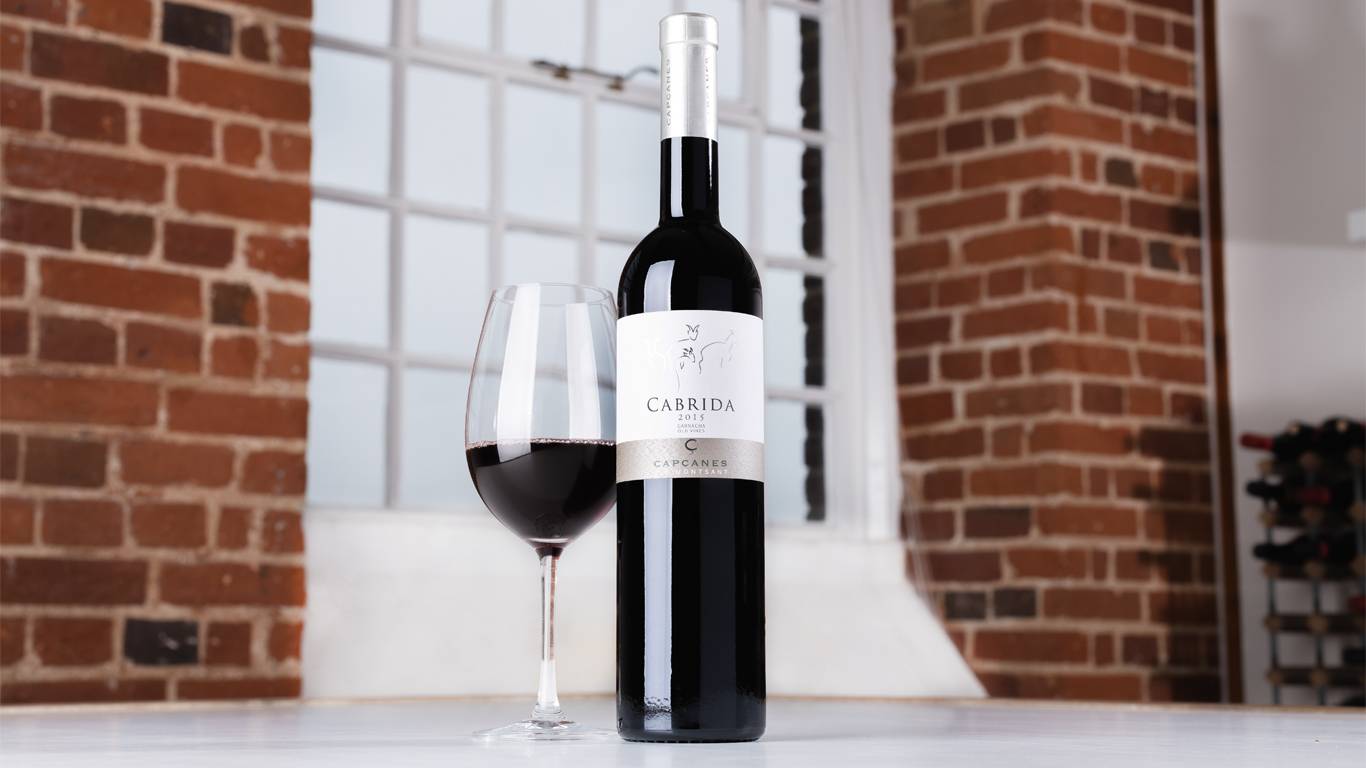 Bottle of Capcanes Cabrida Garnacha 2015 with a glass of red wine on a table in front of a window