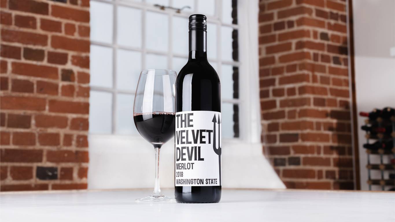 Bottle of Charles Smith Velvet Devil Merlot 2018 with a glass of red wine on a table in front of a window