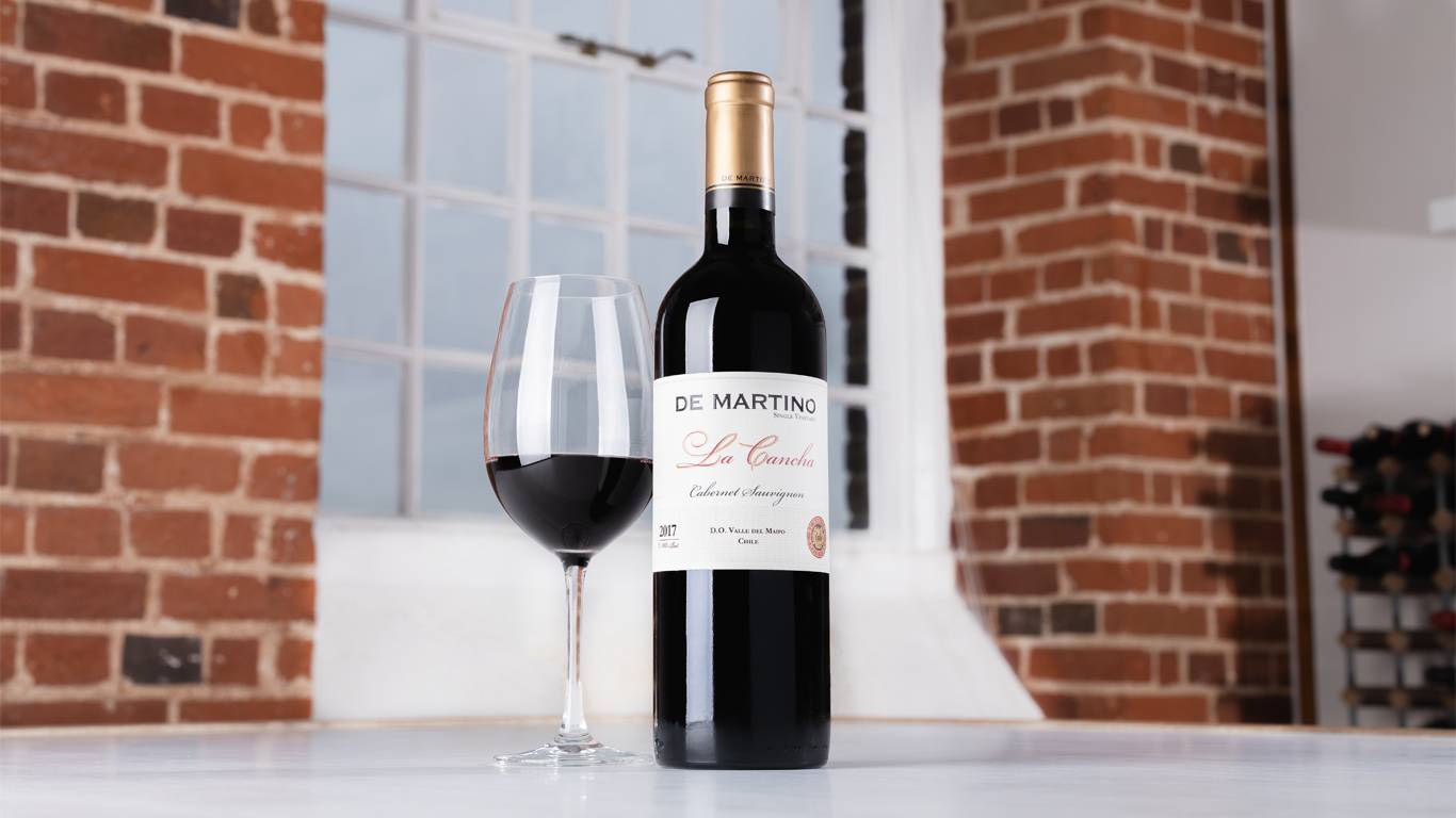 Bottle of De Martino Single Vineyard La Cancha 2017 with a glass of red wine on a table in front of a window