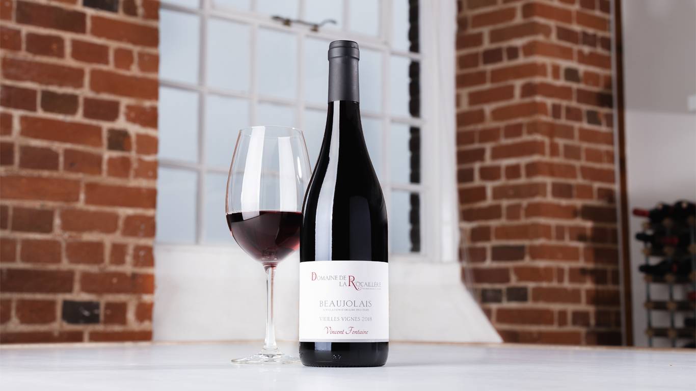 Bottle of Domaine de la Rocaillere Beaujolais Vielles Vignes 2018 with a glass of red wine on a table in front of a window