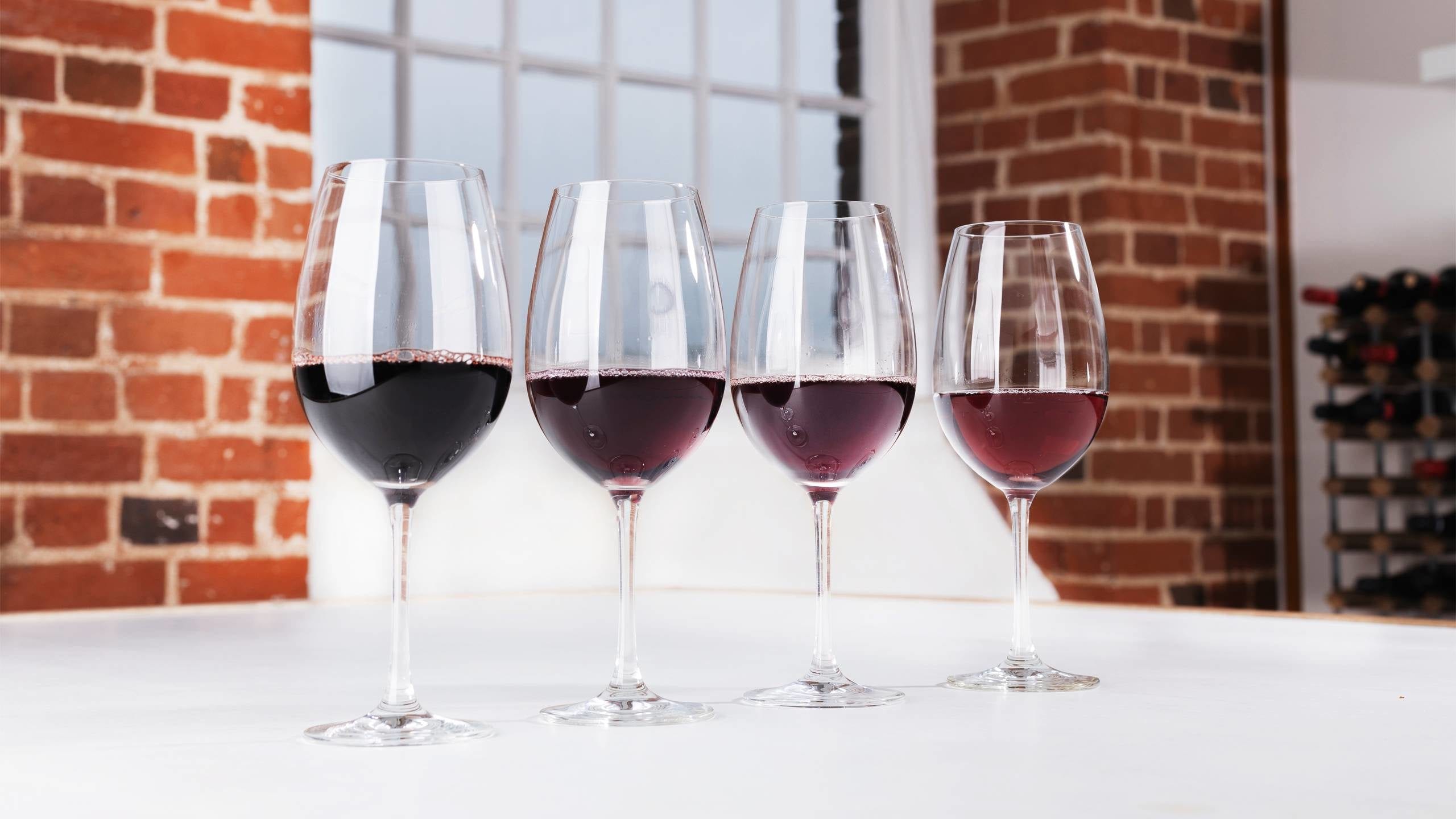 Four glasses of red wine on a table in front of a window and brick wall