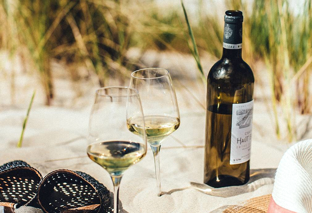 Two glasses of white wine by a bottle in the sand with some sandals and sun hat
