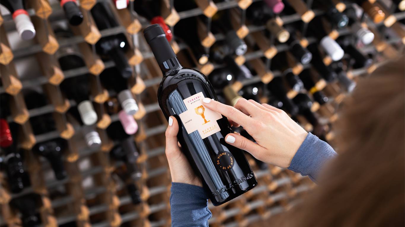 Woman looking at a bottle of red wine in front of a wine rack, and pointing at the front label