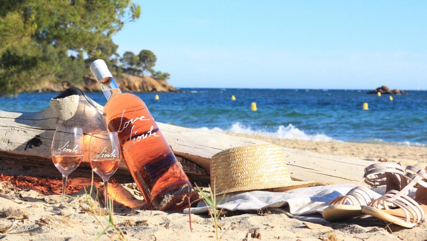 Bottle and two branded glasses of Leoube rose wine, by a log on a beach in Provence
