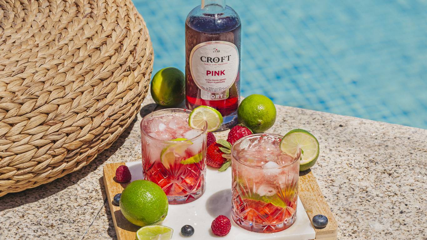 Caipipink Cocktail made with Croft Pink, beside two drinks, sliced lime and berries, on a chopping board beside a swimming pool