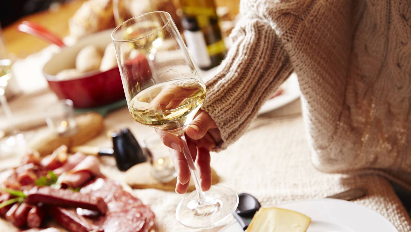 a persons arm in a jumper holding glass of white wine over table with cold meats