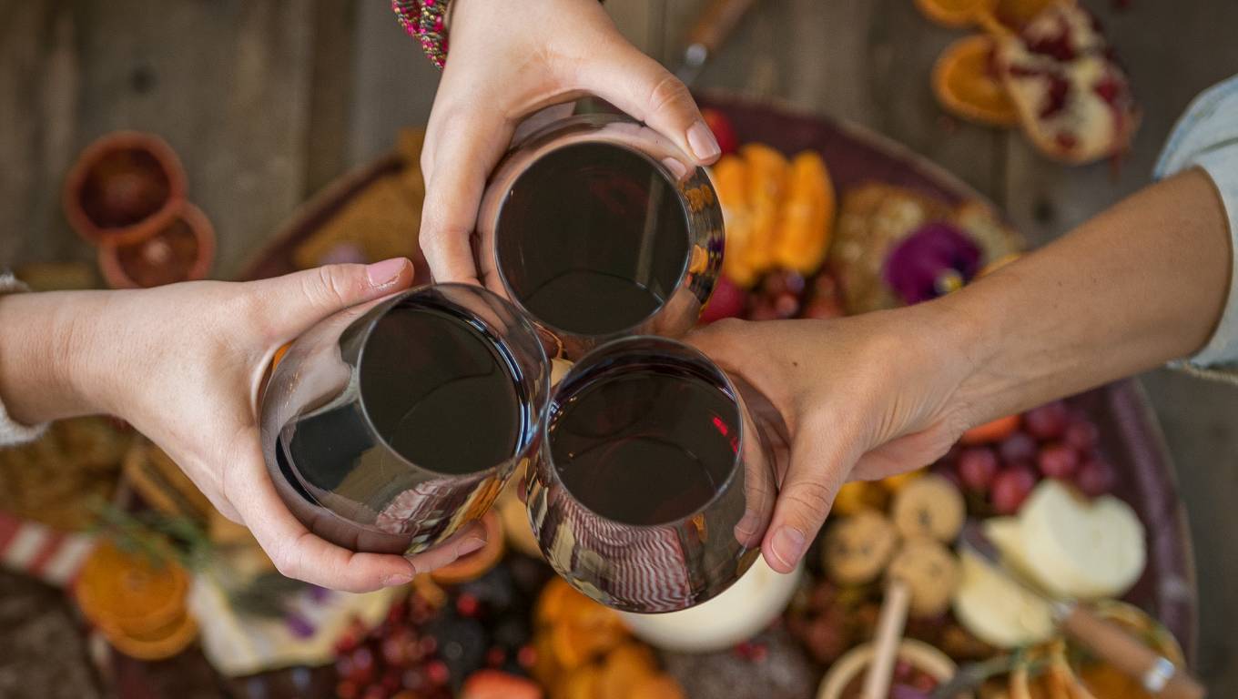 Three people toasting with glasses of red wine over a cheese board