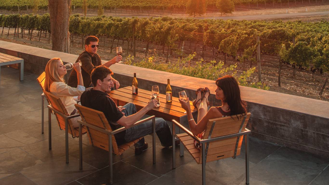 Two men and two women sitting around a table drinking wine overlooking a vineyard at sunset in California