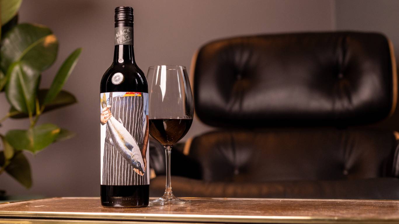 Black Flag Winemakers Limestone Coast Shiraz Cabernet 2018 by a glass of wine on a coffee table by a chair