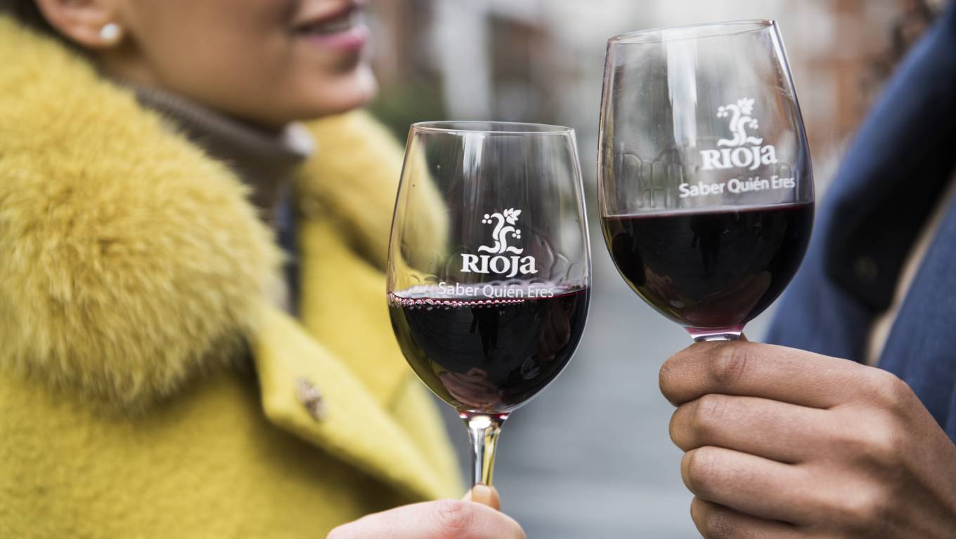 two people holding glasses of red wine with Rioja branded glasses, stood outside in coats