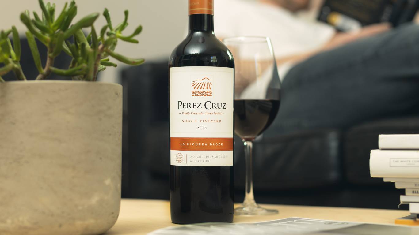 Bottle of Perez Cruz Single Vineyard La Higuera Block on a coffee table with a plant and stack of books, and a man reading a book on the sofa in the background