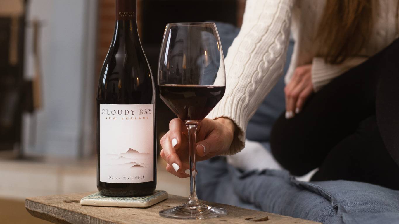 Woman picking up a glass of red wine beside a bottle of Cloudy Bay Pinot Noir 2019 on a coffee table