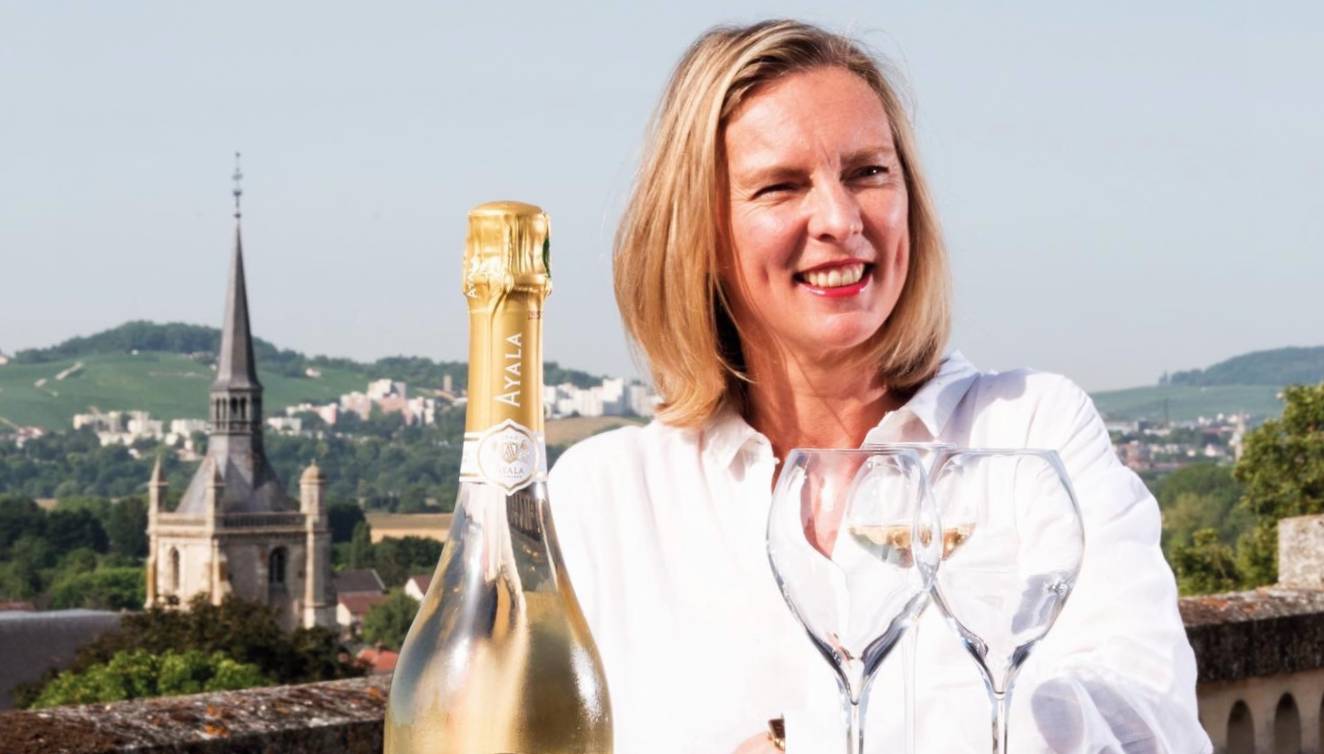 Caroline Latrive of Champagne Ayala sitting outside in the sunshine with stunning views of Champagne behind her and glasses and a bottle of Champagne in front of her