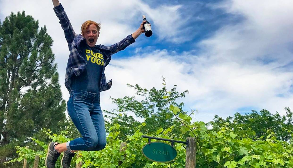 Female winemaker Sara Goedhart of Hedges Family in Washington, jumping high in the air in a vineyard of Syrah while holding a bottle of wine