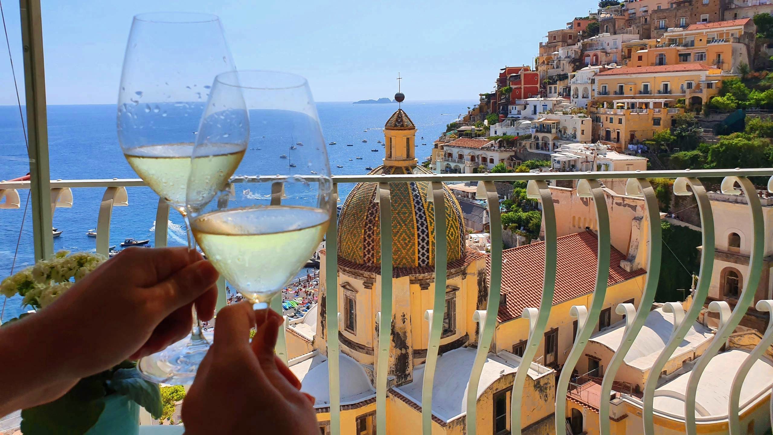 Two hands holding glasses of white wine on a balcony in Italy overlooking Positano town and views of the famous Amalfi Coast