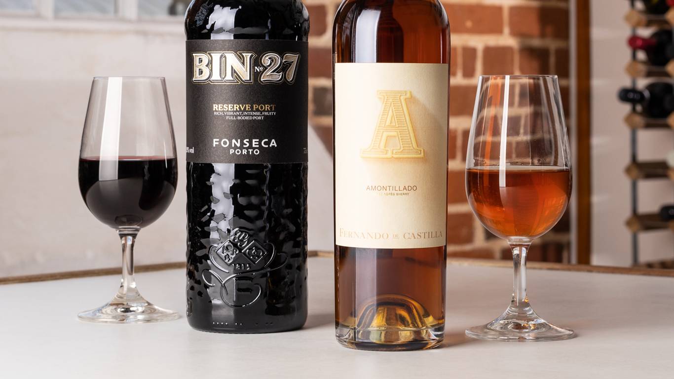 A glass of Port next to a bottle of Port, and a glass of Sherry next to a bottle of Sherry, in front of a window and brick wall