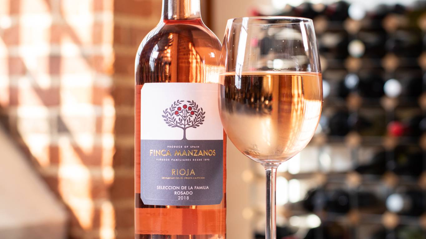 A glass of rose wine next to a bottle of rose wine in front of a brick wall and wine rack