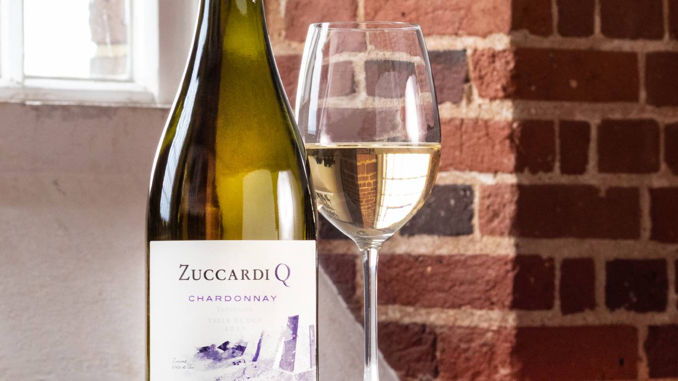 A glass of white wine next to a bottle of Chardonnay in front of a window and brick wall