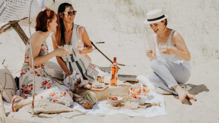 3 women enjoying a glass of rose wine on a beach with food