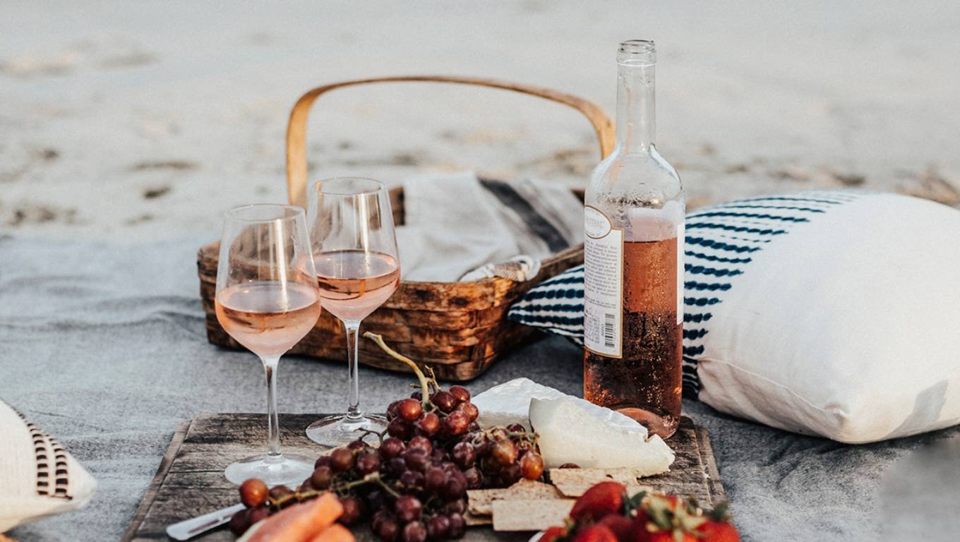 a bottle and 2 glasses of rosé on a blanket on the beach surrounded by food