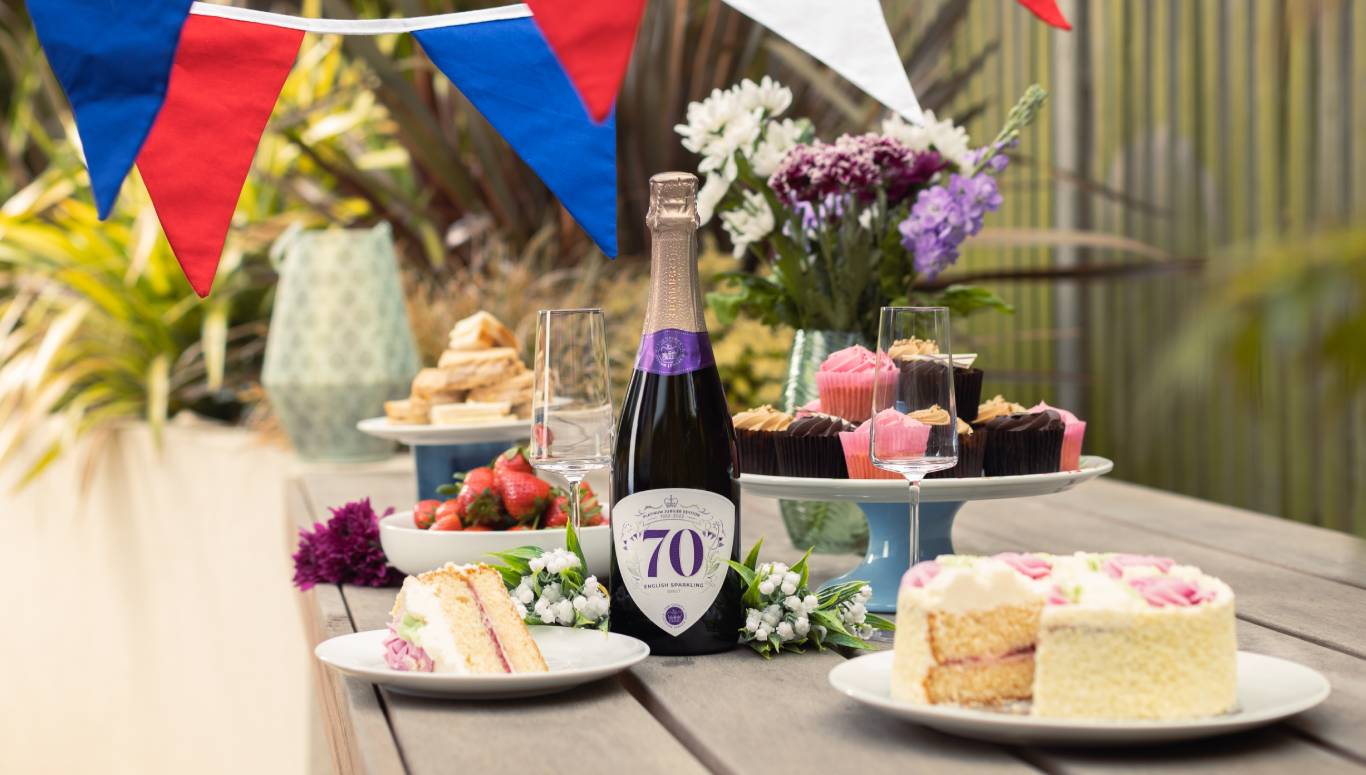 Bottle of commemorative Platinum Jubilee English Sparkling Brut by Virgin Wines on a table outside surrounded by cakes and Jubilee decorations