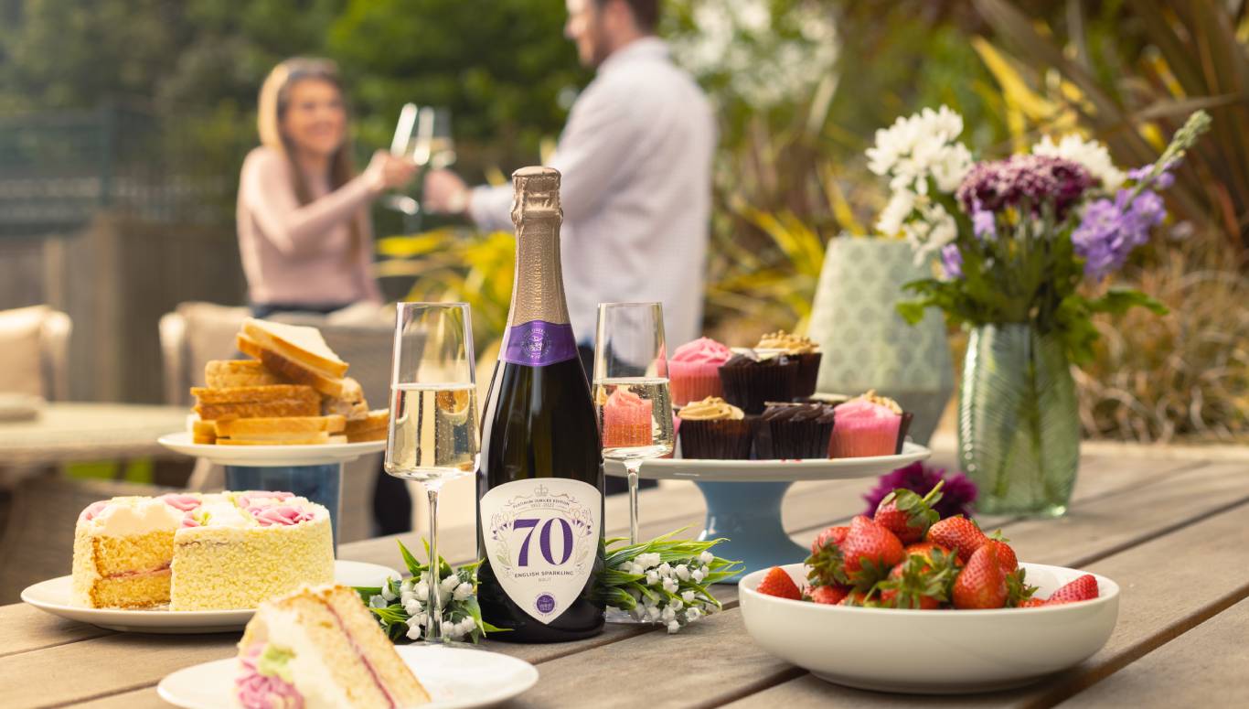 Bottle of commemorative Platinum Jubilee English Sparkling Brut by Virgin Wines on a table outside surrounded by cakes and Jubilee decorations, and two people toasting with Champagne flutes in the background