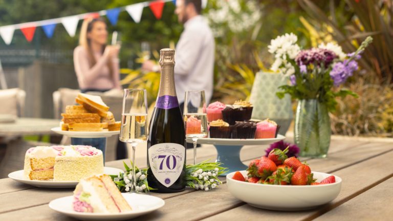Bottle of commemorative Platinum Jubilee English Sparkling Brut by Virgin Wines on a table outside surrounded by cakes and Jubilee decorations, and two people drinking in the background