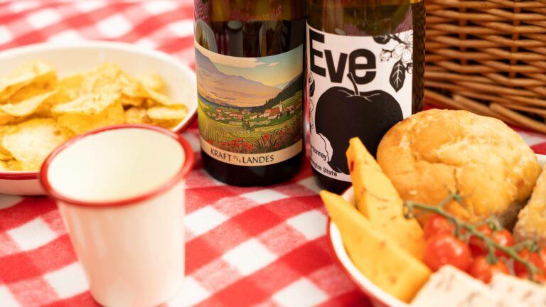 Two bottles of white wine on a red and white gingham picnic blanket with food to represent summer white wines
