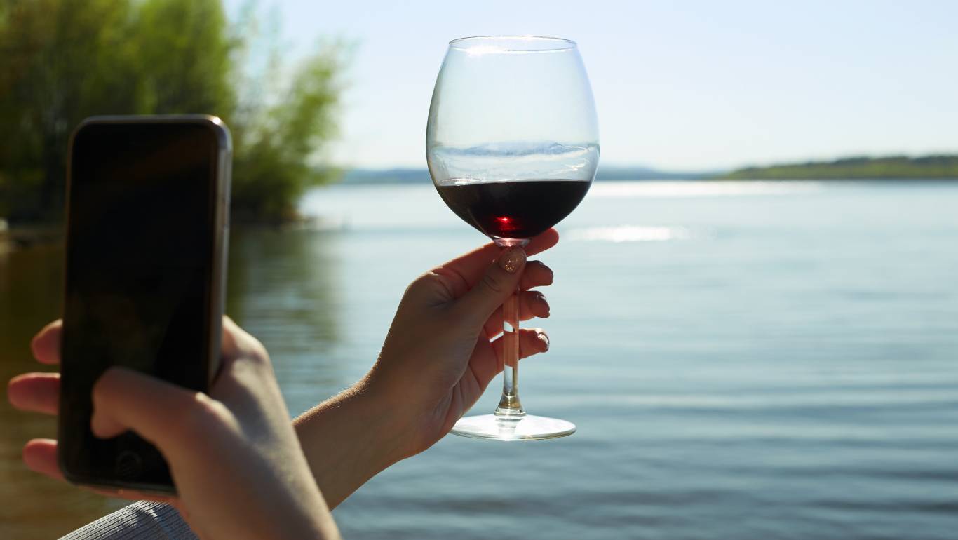 Woman holding up a glass of red wine in front of a lake and taking a photo of the glass and the view with her smartphone