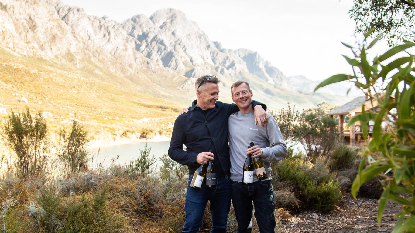 Virgin Wines Wine Buyers, Andrew and Dave, stood in front of Franschhoek Valley in the Western Cape, South Africa, holding bottles of South African wine