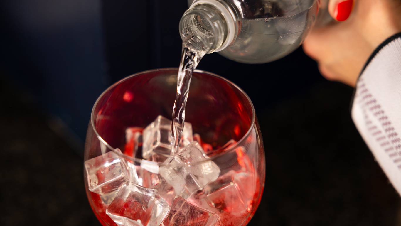 Woman pouring tonic water into a coupe glass filled with red gin and ice cubes