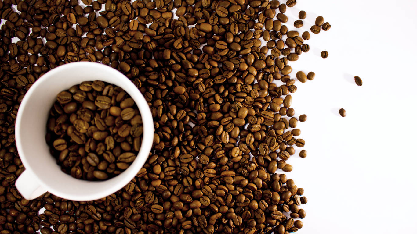 Coffee cup filled with coffee beans and surrounded by lots of coffee beans on a white surface