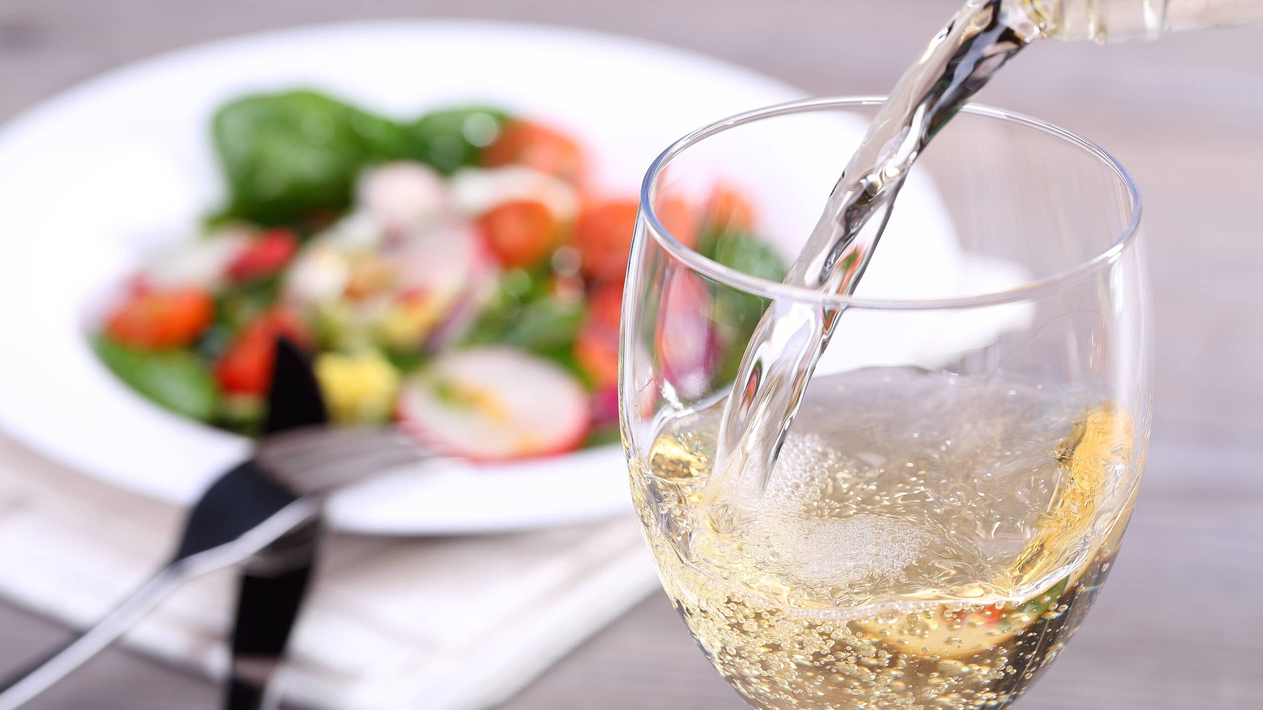 Pouring white wine into glass with plate of salad in the background